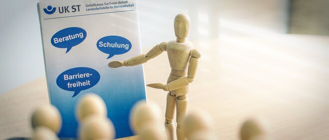 Holzfigur erklärt Plakat der Landesfachstelle mit den Begriffen Beratung, Schulung, Barrierefreiheit - zur Artikelseite Beiträge zum Wissensaufbau 2023