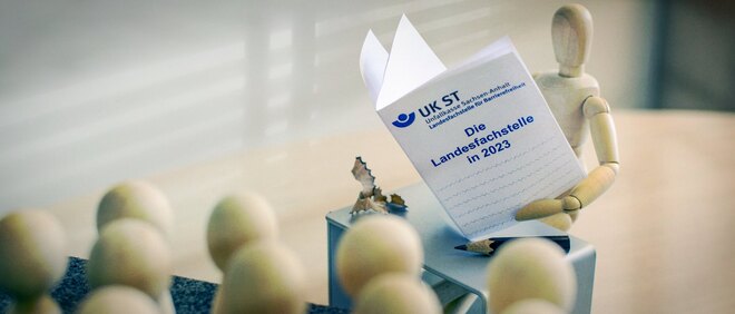 Holzfigur steht an einem Pult und hält Bericht "Die Landesfachstelle in 2023" in der Hand - zur Übersichtsseite die Landesfachstelle 2023